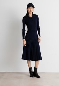 Navy ribbed knit dress with long sleeves and a pleated skirt, paired with black ankle boots and a matching cap. Smooth texture, fitted design.