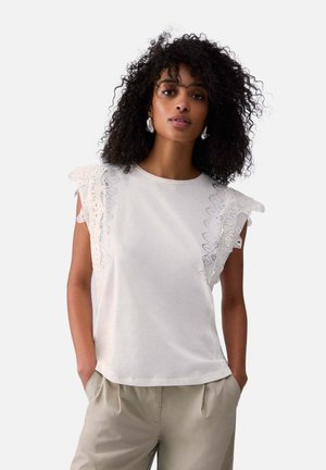 Donna con capelli ricci che indossa un top bianco senza maniche con dettagli in pizzo arricciato e pantaloni beige, in piedi con le mani nelle tasche.