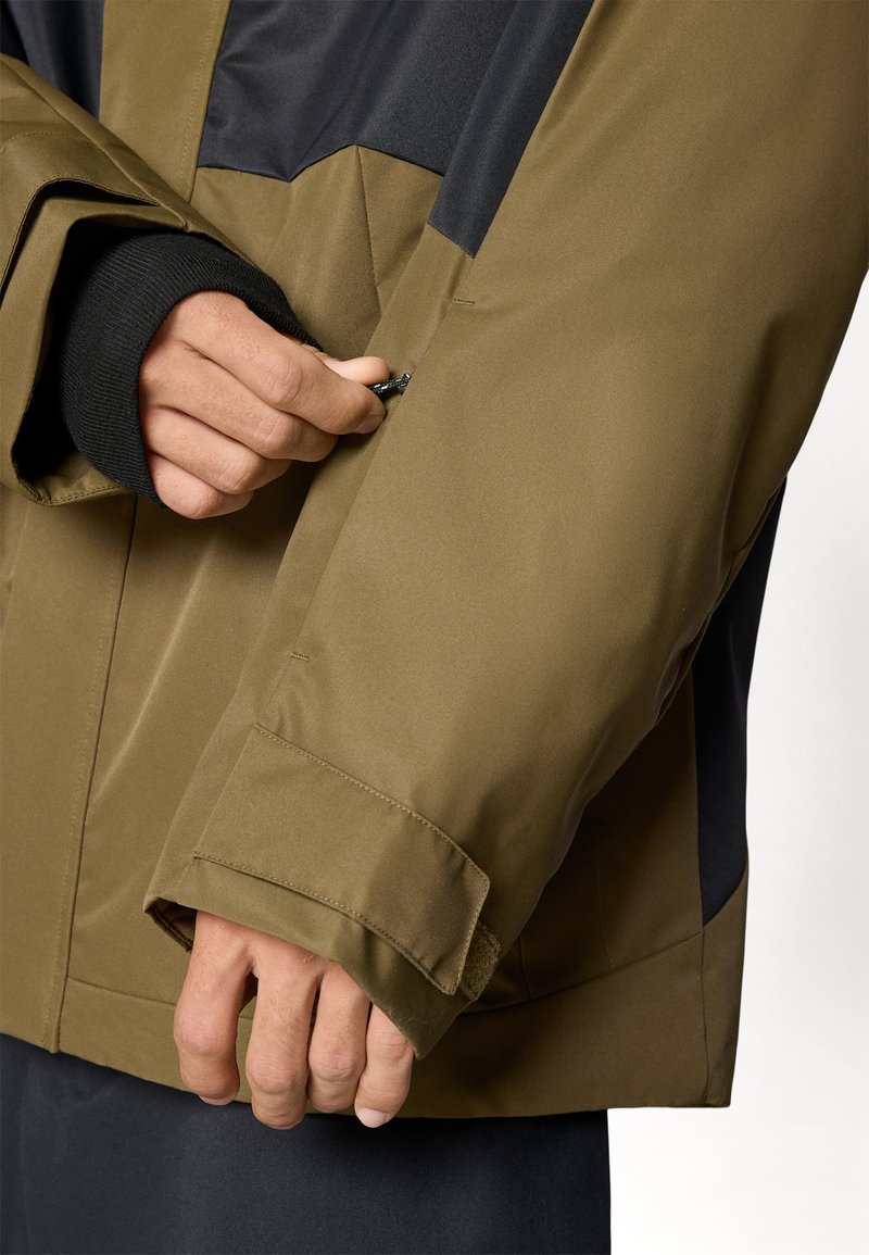 Veste imperméable en vert olive et noir, avec un poignet zippé et un tissu texturé, montrant un gros plan d'une main utilisant la fermeture éclair.