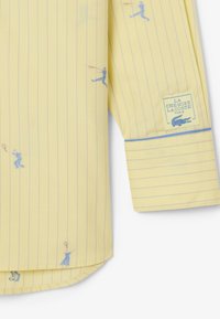 Camicia gialla a maniche lunghe con sottili strisce blu, piccoli motivi di giocatori di tennis e una toppa con il logo La Chemise Lacoste Paris sul polsino.