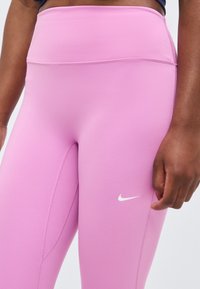 Κοντινή λήψη ατόμου που φοράει ροζ leggings υψηλής μέσης Nike με ορατές ραφές και μικρό λευκό λογότυπο Nike στον μηρό.