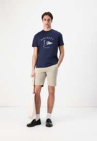 Camiseta de algodón navy con el logo "HACKETT", pantalones cortos beige, zapatos negros y una bolsa de mano color crema. El modelo está de pie frente a un fondo blanco liso.