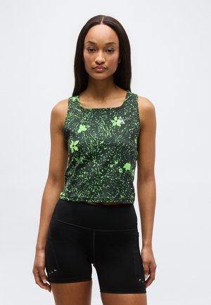 Vrouw draagt een zwart-groen mouwloos cropped top met abstracte patronen en zwarte strakke sportshorts, staand tegen een effen achtergrond.