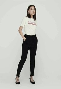 Kvinde med glat sort hår iført hvid "California" T-shirt, høj talje sorte bukser og sorte snørebåndssko, stående med hænderne i lommen.