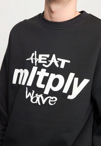 Felpa nera con grande testo grafico bianco: "HEAT mltplly wave," con diverse stili di font e texture sulla parte anteriore.