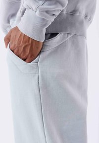 Sudadera gris claro con puños acanalados y un bolsillo lateral. La tela tiene una textura suave y un diseño de corte relajado.