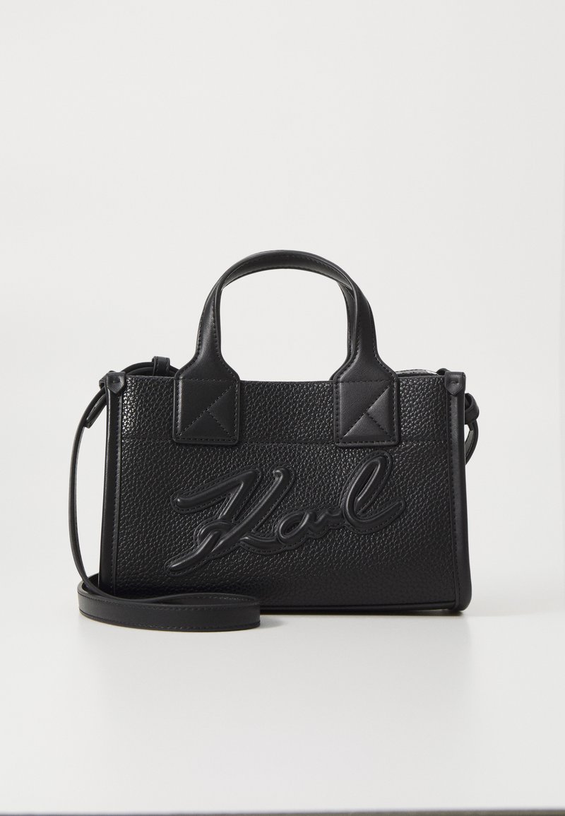 Černá kožená kabelka s texturou, se dvěma vrchními rukojeťmi a odnímatelným ramenním popruhem. Na přední straně je vyvýšené logo "Karl".