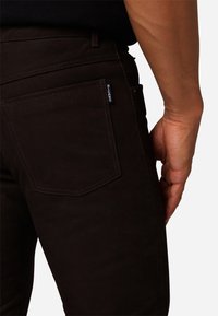Donkerbruine broek met een achterzak met zichtbare stiksels en een merketiket. De stof heeft een gladde, licht gestructureerde uitstraling.