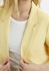 Yellow tailored blazer made of smooth fabric, featuring notched lapels and a slightly structured fit, paired with a white ribbed top.
