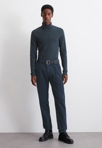 Donkergroene coltrui, aangesloten fit, gecombineerd met marineblauwe, op maat gemaakte broeken. Zwarte riemdetails en stevige zwarte veterschoenen. Eenvoudig, modern ontwerp.