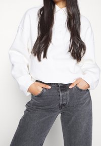 Vit långärmad polo med knappkrage och avslappnad passform, tillsammans med högmidjade mörkgrå jeans med sidofickor.