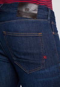 Pantalones vaqueros de mezclilla azul oscuro con una etiqueta de cuero y costuras en contraste de color naranja. Presentan un bolsillo trasero con costuras decorativas en rojo y azul marino.