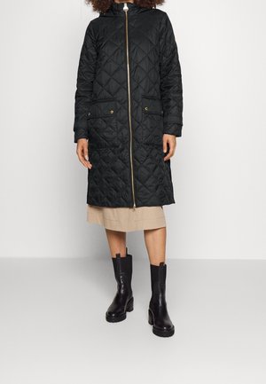 Femme portant un long manteau matelassé noir avec une fermeture éclair dorée, une jupe beige et des bottines noires sur fond blanc uni.