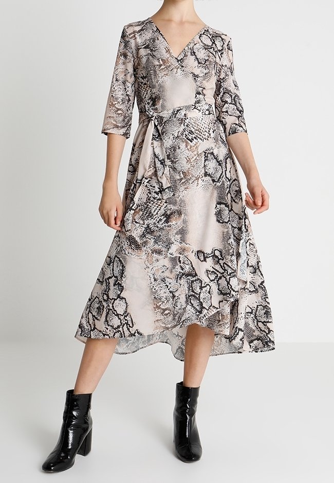 Femme vêtue d'une robe portefeuille à imprimé serpent avec des manches trois-quarts et des bottines noires à talons, debout devant un fond uni.
