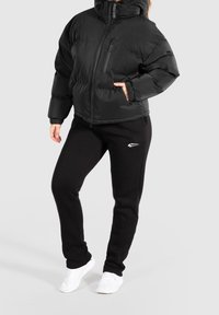 Giacca a vento nera con collo alto, chiusura con zip e tasche laterali; abbinata a pantaloni della tuta neri con logo bianco; sneaker bianche.