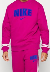 Sudadera fucsia con "NIKE" en azul a lo largo del pecho, puños acanalados con rayas blancas y pantalones a juego; material de algodón con una textura suave.