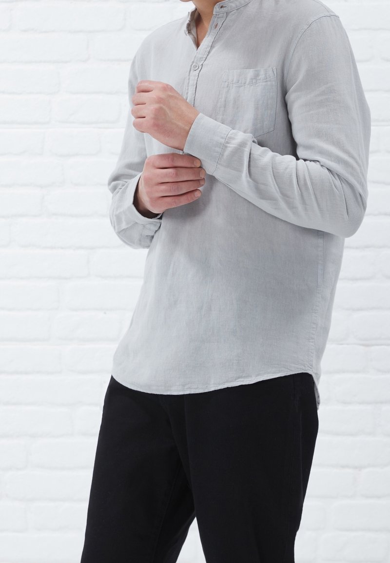 Lichtgrijze langemouwen shirt met een henley-halslijn, een borstzak en een relaxte pasvorm, gecombineerd met zwarte broek voor een witte bakstenen achtergrond.