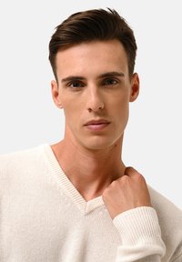 Weißer, gestrickter Pullover mit V-Ausschnitt und gerippten Bündchen, getragen von einer Person mit kurzem Haar und neutralem Ausdruck.