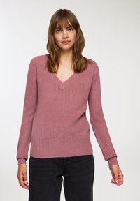 Sweter w kształcie litery V w miękkim, teksturowanym, przydymionym różowym splocie. Posiada długie rękawy, ściągacze na mankietach i u dołu oraz luźny krój. W zestawie z czarnymi dżinsami.