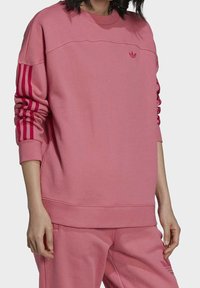 Rosa sweatshirt med ribbade muddarna och fållen, med tre vertikala ränder i en mörkare rosa nyans på ärmarna och en liten logotyp på bröstet.