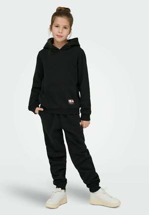 Young girl standing wearing black hoodie and matching joggers, paired with white sneakers, on plain light background.