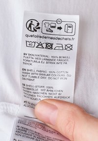 Vaatehoito-ohje valkoisella kankaalla, jossa on pesuohjeet useilla kielillä. Materiaali: 100 % puuvillaa. Sisältää pesusymboleita.