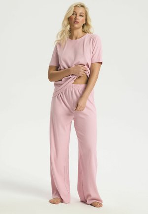 Žena so svetlými vlasmi, oblečená v voľnom ružovom rebrovanom tričku s krátkym rukávom a zodpovedajúcimi širokými nohavicami, stojaca bosá na bielom pozadí.