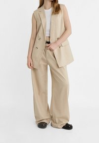 Beżowa, bezrękawnikowa kamizelka na miarę z dwurzędowymi guzikami, w zestawieniu z szerokimi beżowymi spodniami. Gładka tkanina, minimalistyczny design i czarny pasek.
