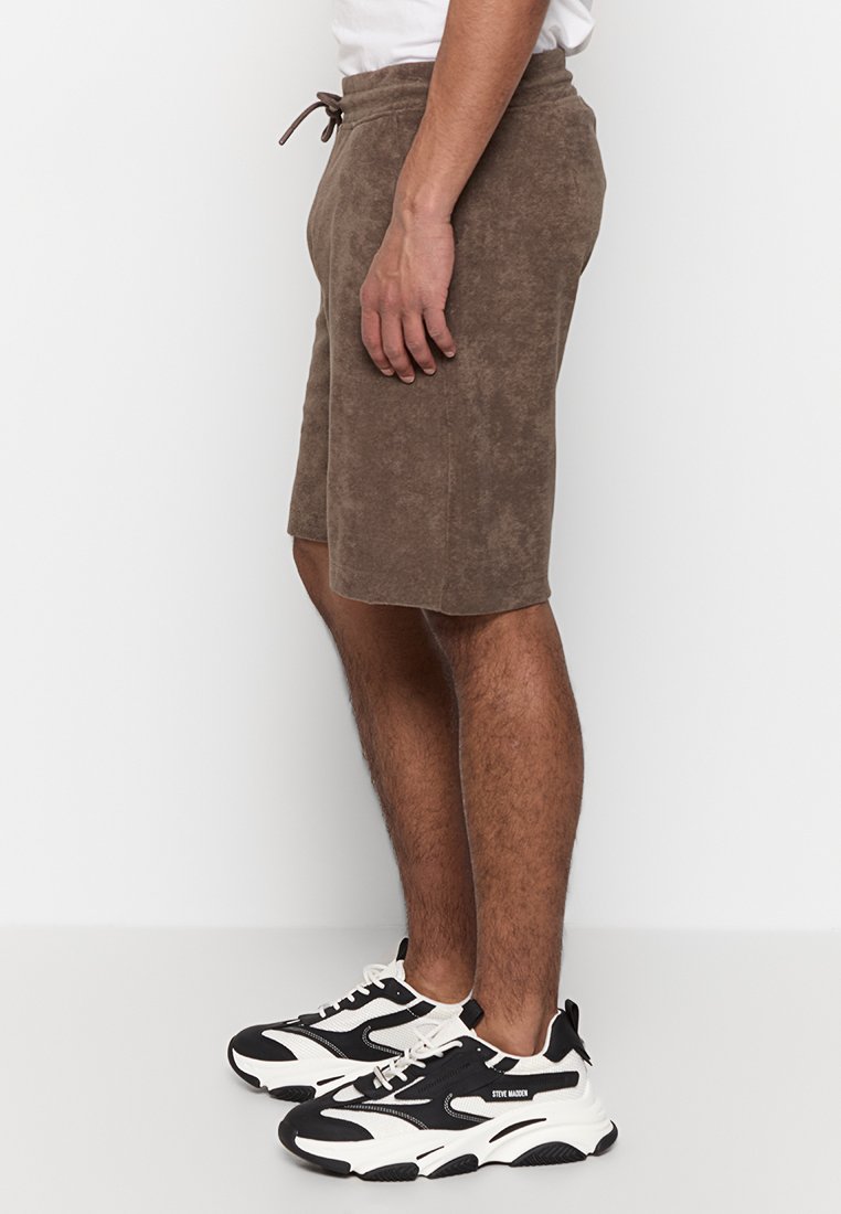 Mann in braunen knielangen Shorts und schwarz-weißen Turnschuhen, der vor einem schlichten weißen Hintergrund steht.