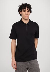 Svart kortermet polo med glidelås i halsen, laget av glatt stoff. Modellen har på seg bukser i grått med lett tekstur.