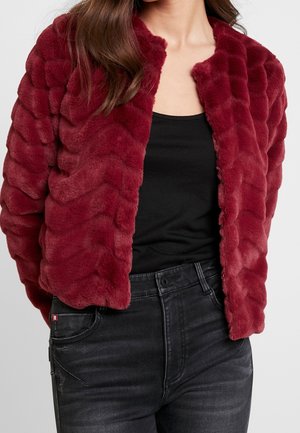 Frau trägt eine strukturierte, tiefrote Kunstpelzjacke über einem schwarzen Oberteil und hoch taillierte dunkelgraue Jeans, die am Hals ausgeschnitten sind.