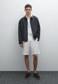 Veste oversized grise, chemise blanche et shorts clairs. Chaussures en daim marron avec une semelle en caoutchouc tan. Tissus lisses, design moderne et couleurs neutres.