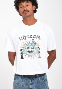 Witte katoenen t-shirt met een kleurrijke grafische print van een lachend gezicht, puzzelstukken en speelse figuren.