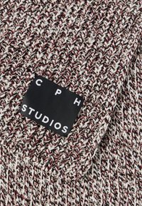 Pletená čiapka z bielej, červenej a čiernej priadze, s štítkom „CPH STUDIOS“ bielym písmom na čiernom pozadí. Viditeľný textúrovaný vzor.