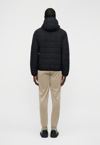 Μαύρο φουσκωτό μπουφάν με καπιτονέ σχέδιο και κουκούλα, με οριζόντιες ραφές και άνετη εφαρμογή, φορεμένο πάνω από μπεζ παντελόνι και μαύρα παπούτσια.