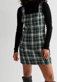 Abito a quadri verde e bianco con scollatura quadrata e forma attillata; abbinato a un dolcevita nero a maniche lunghe sotto.