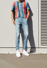 Hombre vestido con una camiseta a rayas azul, roja y blanca, jeans azul claro, calcetines blancos y zapatillas blancas, de pie contra una pared gris.