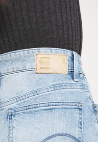 Ljusblå denimjeans har en läderlabel med "RAW" ingraverat, synlig sömnad och en bakficka med rundad form.