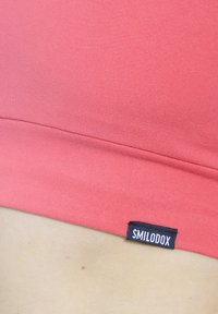 Korallenrotes Sportoberteil aus glattem Stoff, mit einfarbigem, nahtlosem Design und einem kleinen schwarzen Etikett mit der Aufschrift "SMILODOX".