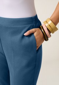 Lichtblauwe op maat gemaakte broeken met een gladde textuur en een elastische tailleband, gecombineerd met meerdere gekleurde armbanden om de pols.