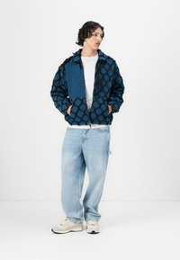 Blue patterned jacket with a fuzzy texture, zippered front, and two pockets, paired with light denim jeans and beige sneakers.