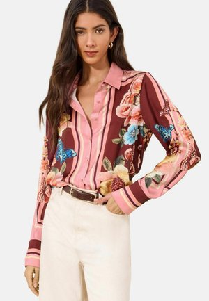 Woman wearing a long-sleeve button-up shirt with pink and brown stripes, floral and butterfly patterns, tucked into light beige pants with a belt.