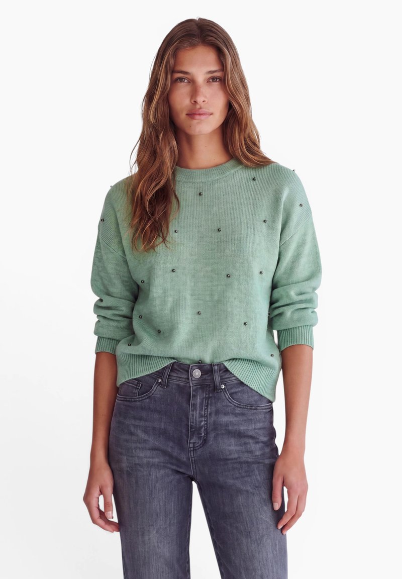 Femme aux longs cheveux ondulés portant un pull vert clair avec de petites décorations et un jean taille haute gris foncé, debout sur un fond blanc.