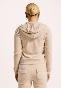 Beige hoodie met een zachte textuur, aansluitend design en een capuchon. Gecombineerd met bijpassende broeken met twee achterzakken en gouden accenten.