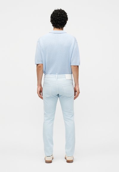 Άνδρας με σγουρά μαλλιά στέκεται με την πλάτη, φοράει γαλάζιο κοντομάνικο πουκάμισο, ανοιχτόχρωμα τζιν και μπεζ παπούτσια σε λευκό φόντο.