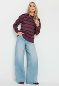 Felpa a righe bordeaux e nere con maniche lunghe, abbinata a jeans a gamba larga azzurri, che mostrano un fit rilassato e uno stile casual.