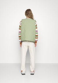 Kvadret grön väst med rund hals, ryggventiler och stora ärmhål, bärs över en multi-randig tröja och matchas med off-white byxor.