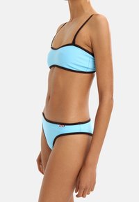 Jasnoniebieski zestaw bikini z bandeau w brązowym wykończeniu oraz dopasowanymi niskimi majtkami, przyozdobiony małym detalem w kolorze czerwono-białym.