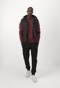 Chaleco tipo puffer con capucha en negro sobre un sudadera burdeos, combinado con pantalones joggers negros y zapatillas blancas. Texturas suaves y diseño minimalista.
