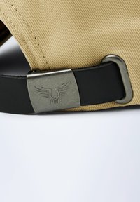 Casquette en tissu beige avec une sangle en cuir noir et une boucle métallique présentant un motif d'aile en relief, mettant en valeur une surface texturée.